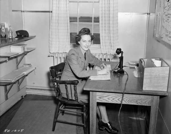 Photographie en noir et blanc – Une femme en tenue militaire est assise à un pupitre en bois dont la surface est couverte de feuilles. On aperçoit une petite étagère et un microphone ainsi que des tablettes et, à gauche, une fenêtre.